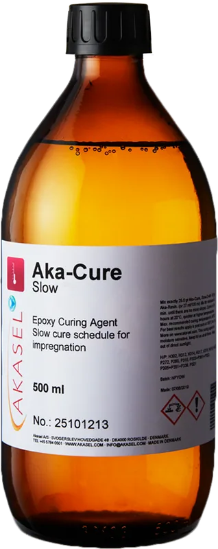 Akasel Aka-Cure Slow: En brun glasflaska med gul skruvkork. Etiketten visar Akasel-logotypen, produktnamnet "Aka-Cure Slow" och "Epoxy Curing Agent." Ytterligare text inkluderar "Slow cure schedule for impregnation," volymangivelsen "500 ml" och specifika blandningsinstruktioner. Säkerhets- och användningsinstruktioner samt varningssymboler finns också på etiketten. Flaskan innehåller en klar vätska, speciellt utvecklad för metallografiska applikationer, idealisk för förberedelse av värmekänsliga prover och impregnering.